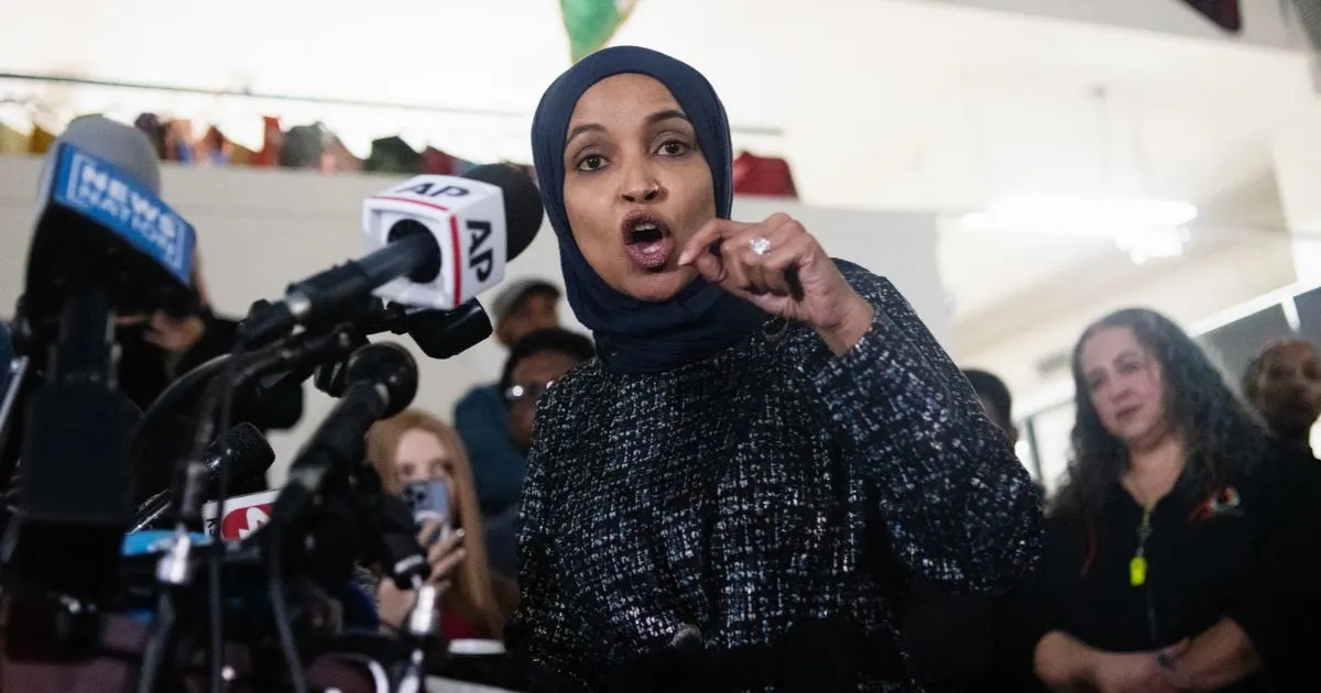 Woman in headscarf and dark jacket speaking at a press microphone cluster, gesturing, with onlookers behind.