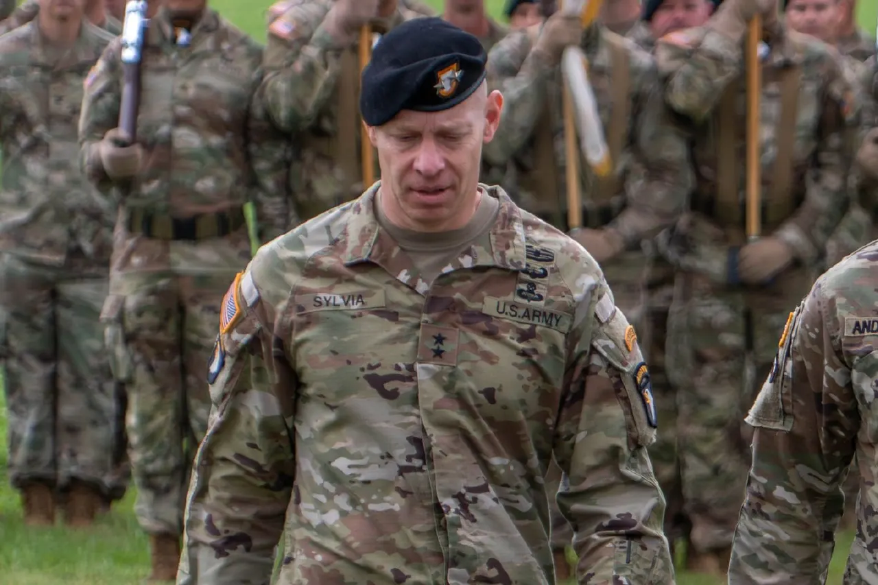 Uniformed U.S. Army officer in black beret, eyes lowered, at a formation ceremony with soldiers behind.