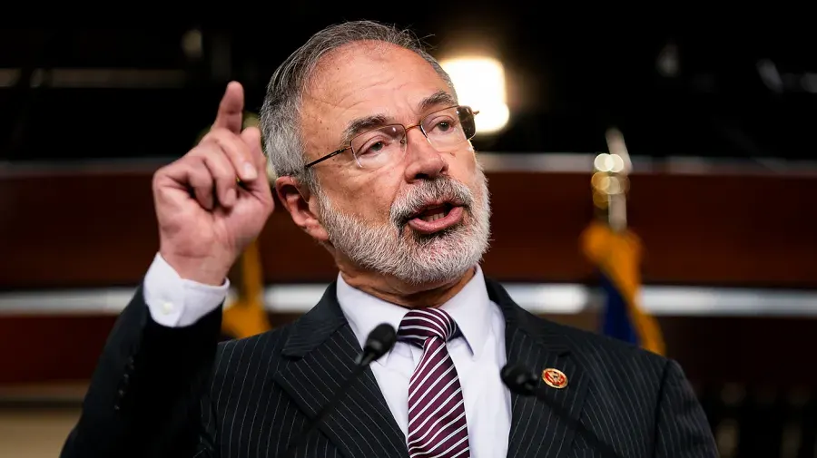 Bearded man in glasses and pinstriped suit pointing upward while speaking at a press microphone.