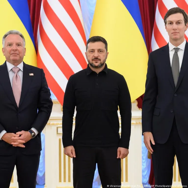Three men in dark suits and a black shirt stand before Ukrainian and American flags in a formal room.