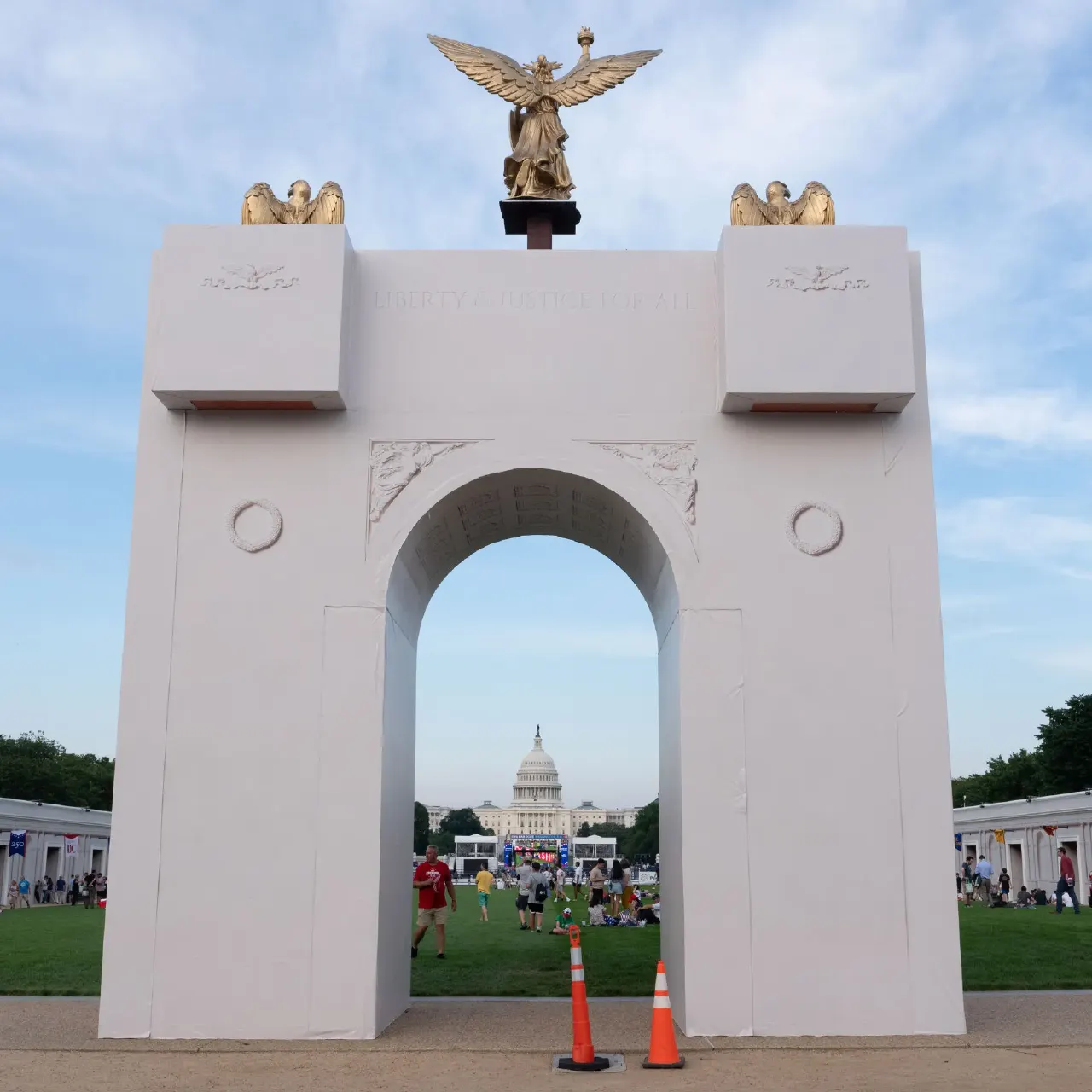 White mock-up triumphal arch framing the U.S. Capitol dome, with people on the lawn