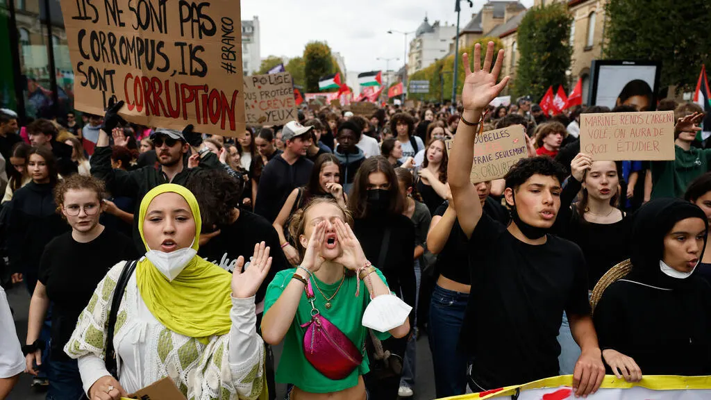 Large street protest with hand-painted signs, young marchers chanting, raised hands, and red and Palestinian flags.