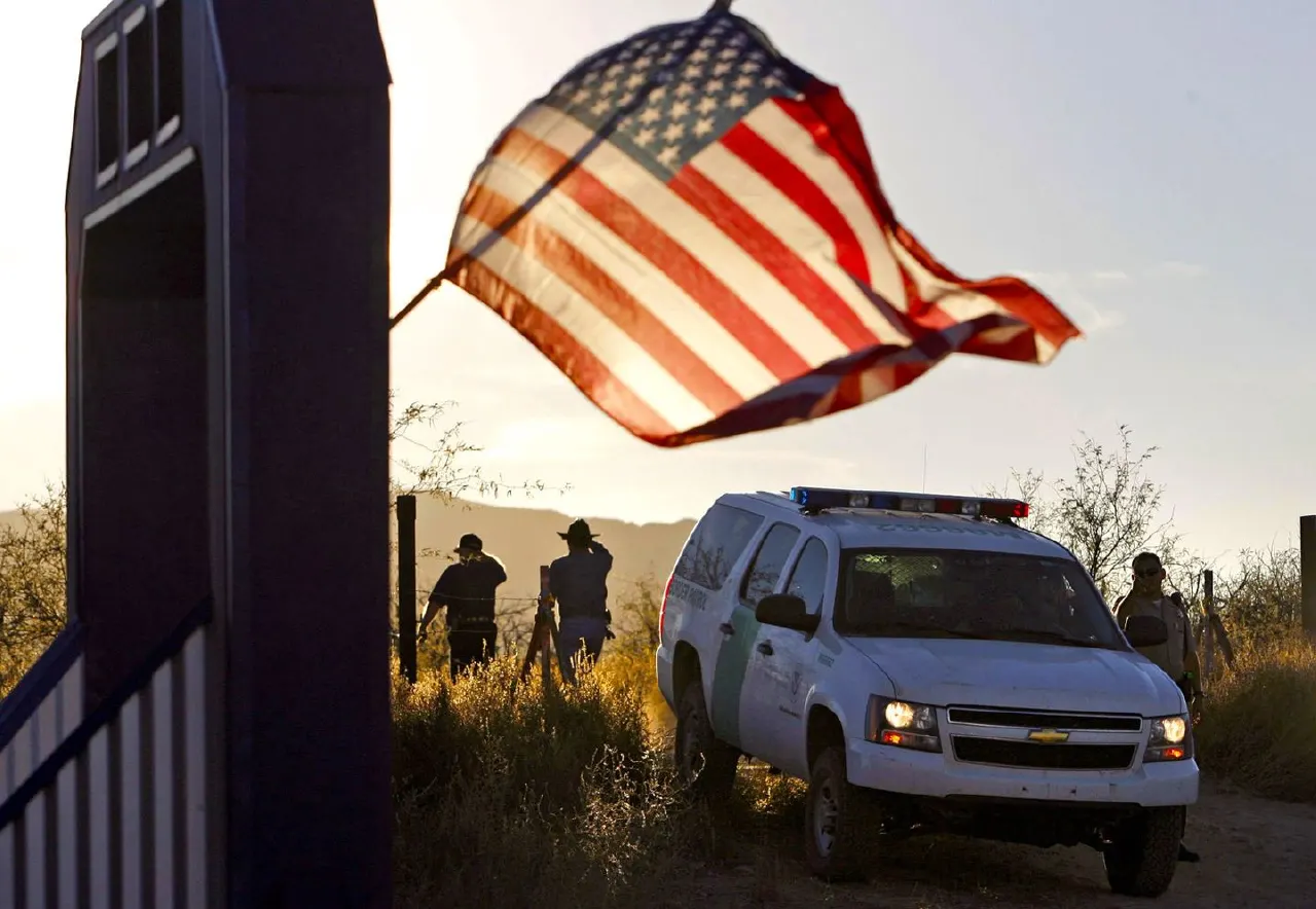 Border agents and white patrol SUV by border fence at sunset, American flag waving overhead.