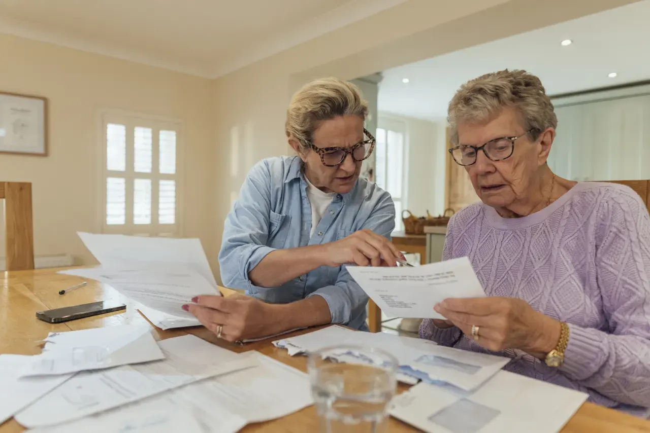 Two older women at a wooden table reviewing paperwork and bills together, one pointing at a document.