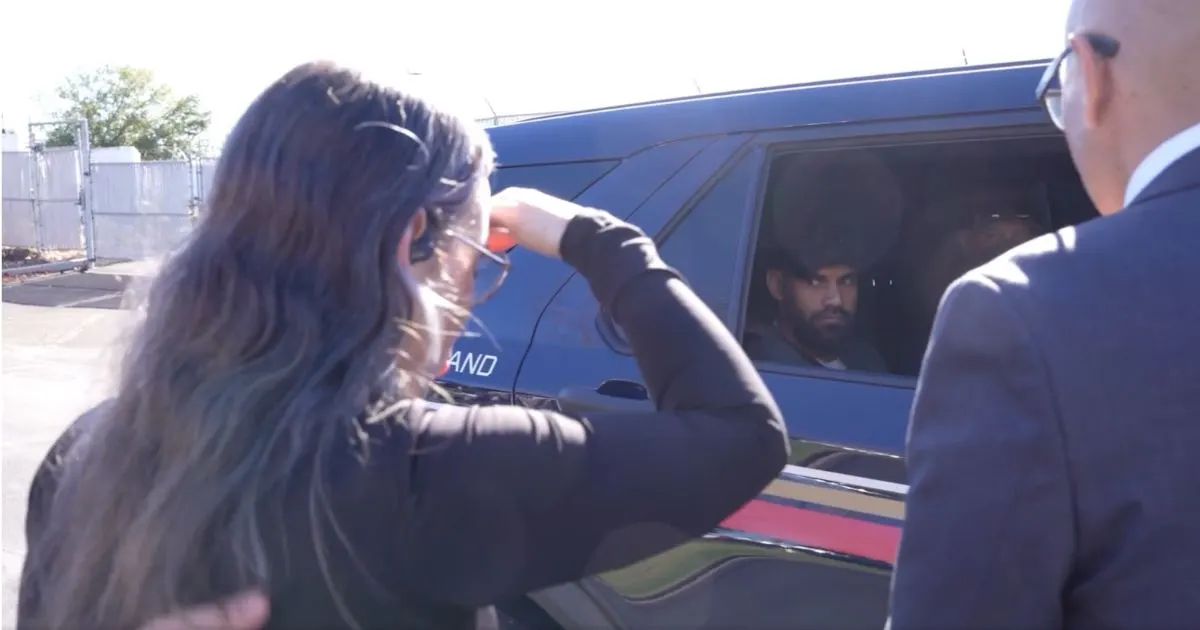 Woman gestures toward a dark SUV where a man sits in the rear window, another man in suit beside.
