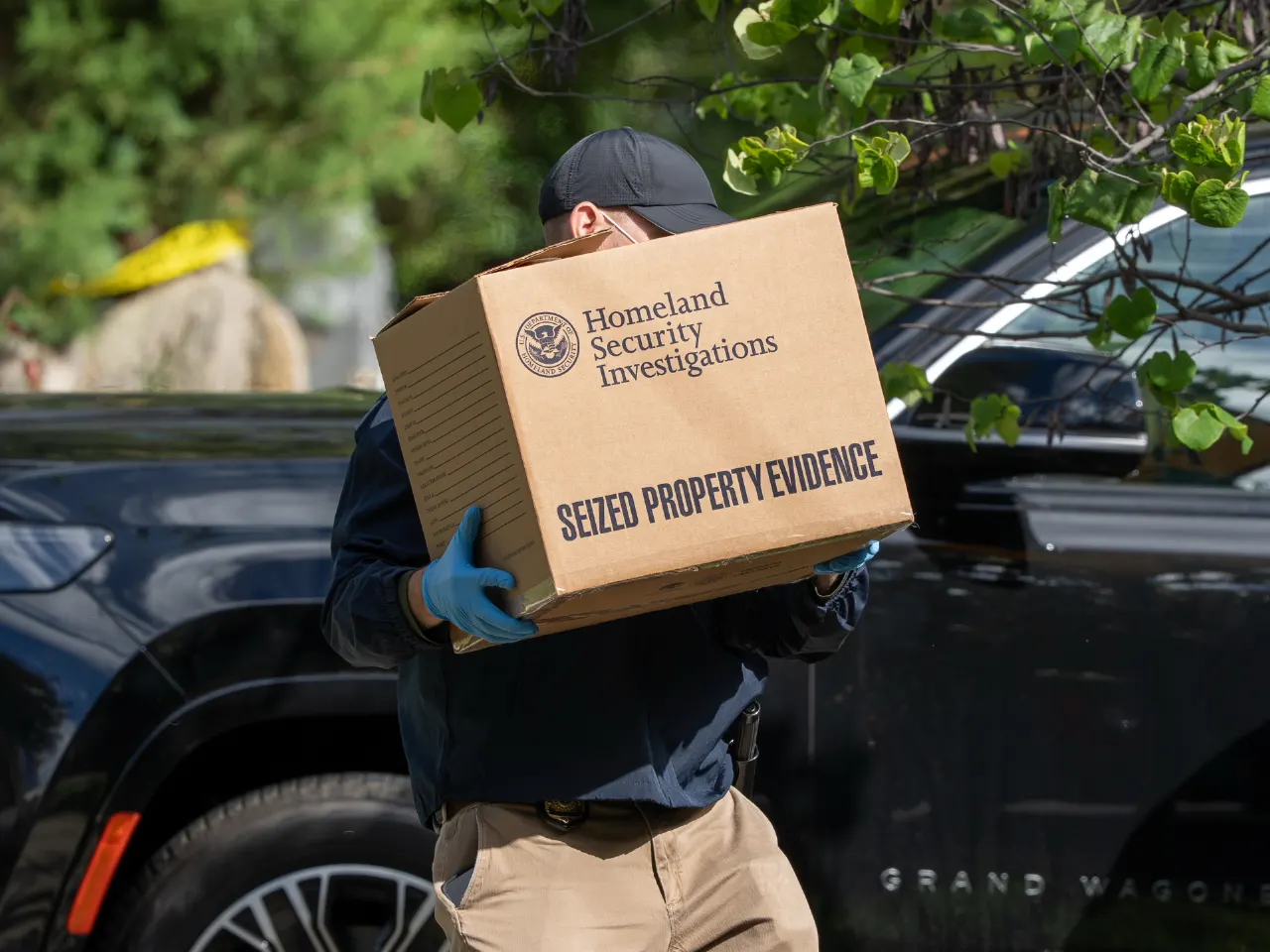 Person in cap carries box marked Homeland Security Investigations seized property evidence past black SUV.