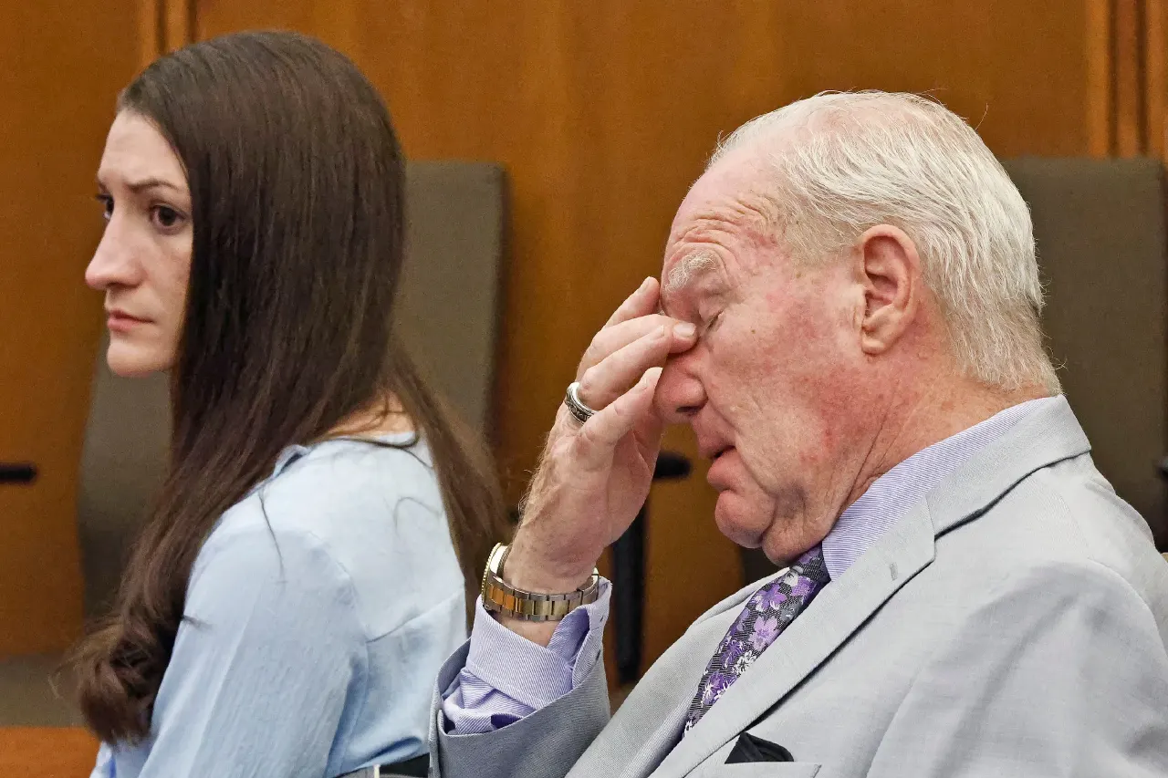 Older man in grey suit rubs his forehead while a woman with long brown hair sits beside him in a courtroom.