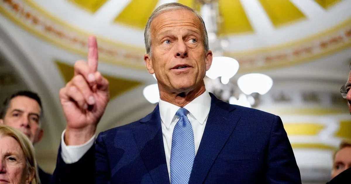 Man in navy suit pointing upward while speaking, with people and gilded ceiling behind him.