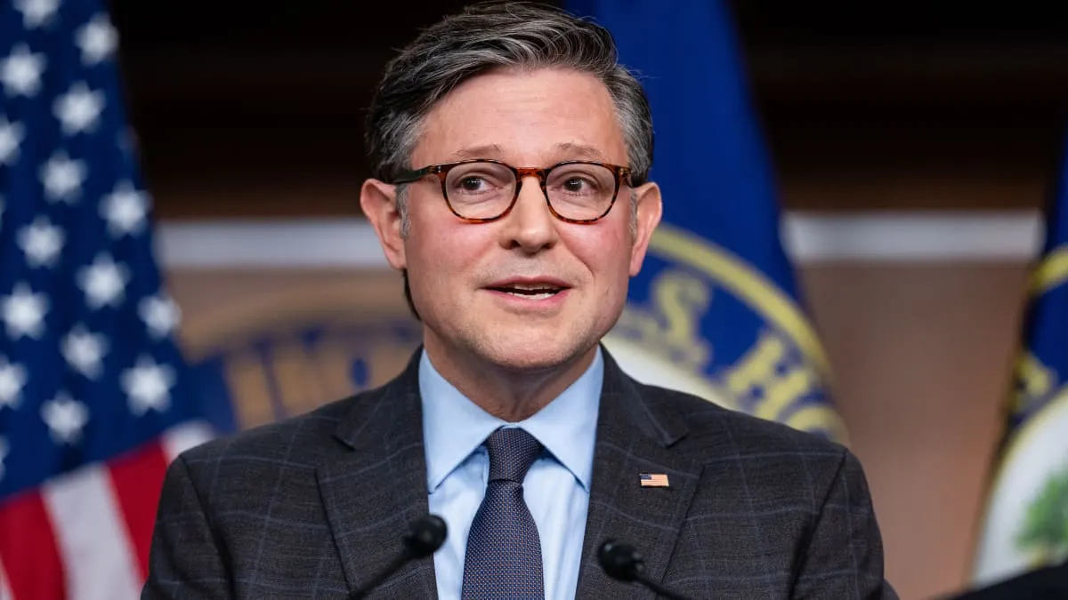 Man in glasses and gray suit speaking at a press event before American and seal flags.