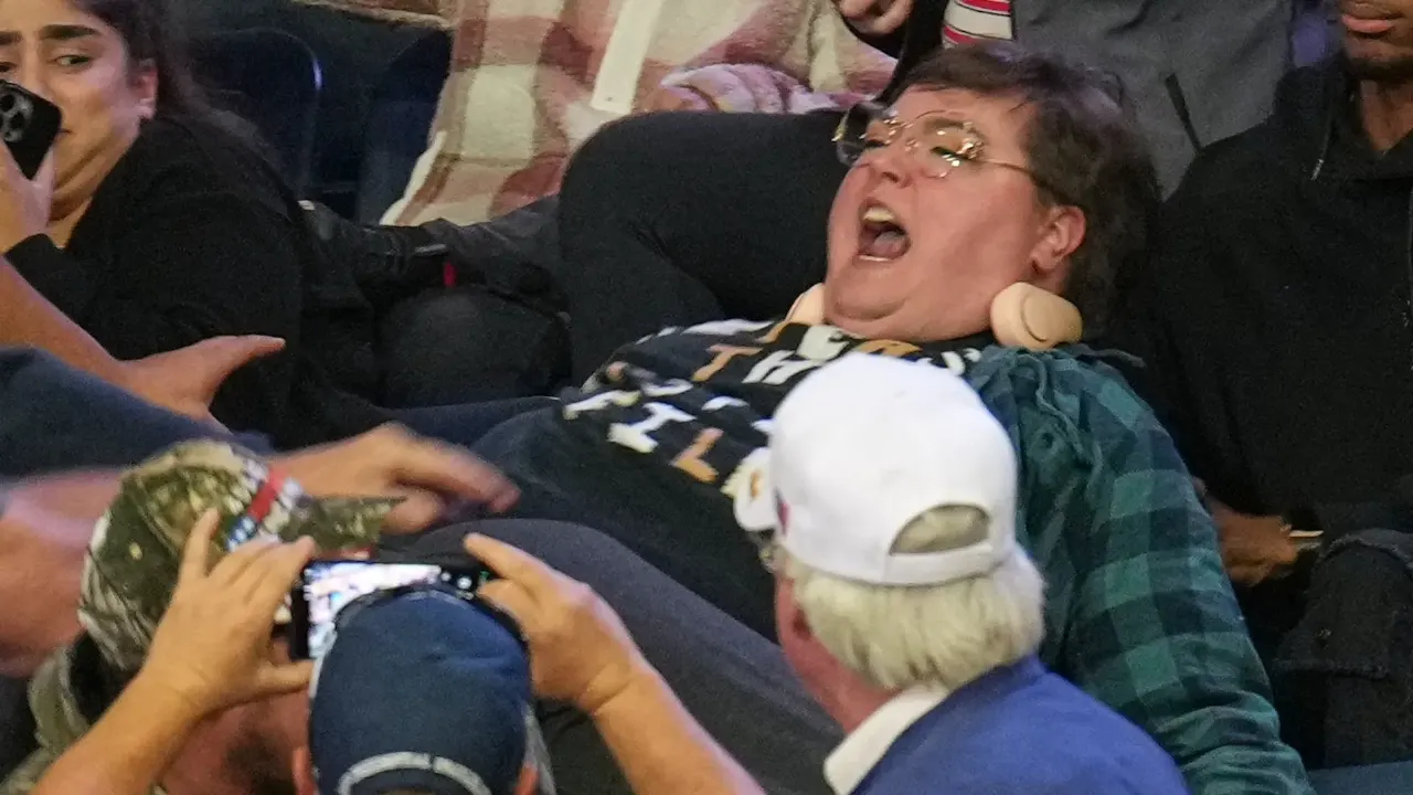 Woman with glasses shouting while seated in crowd, people around her holding up phones.