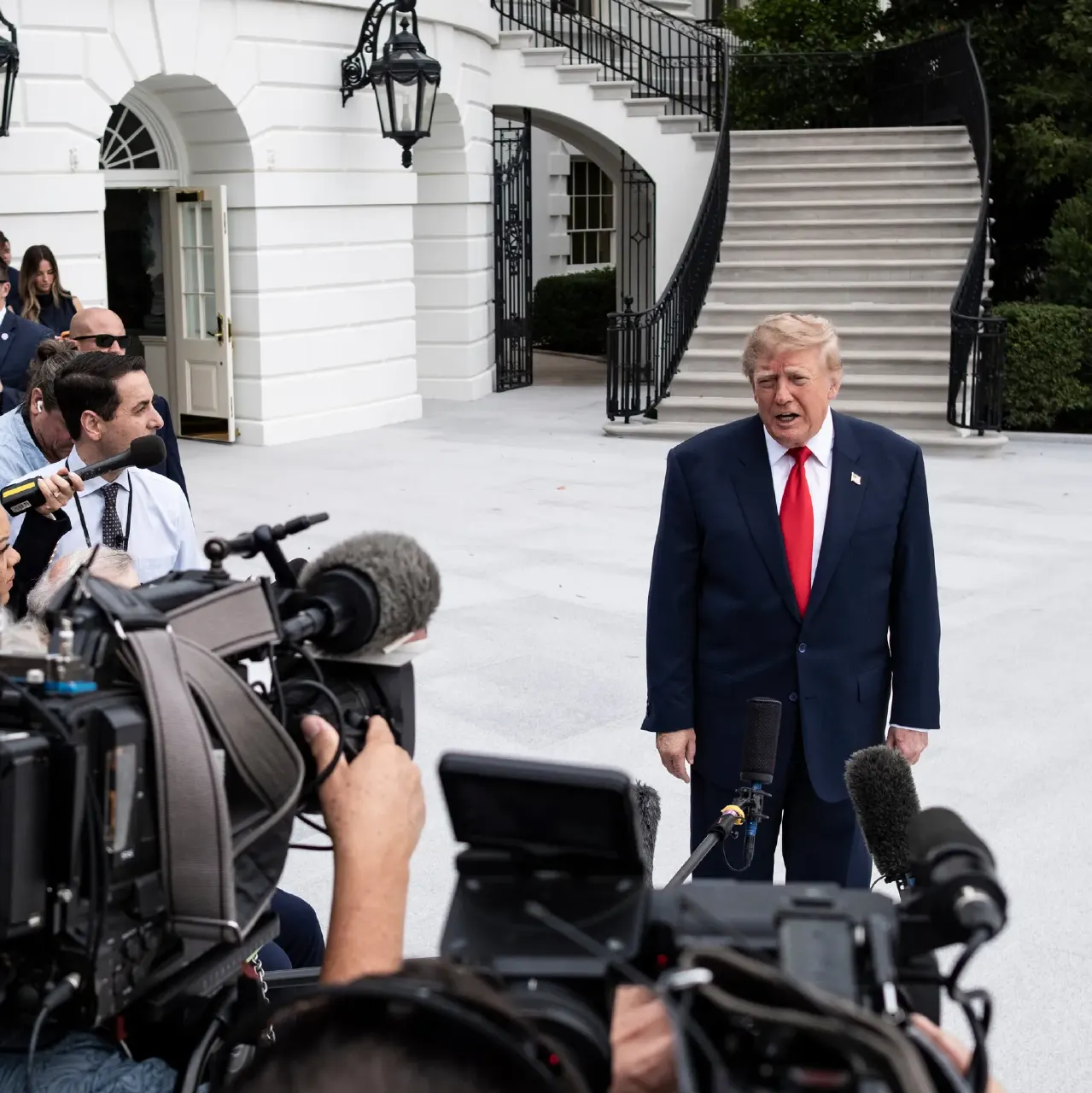 A man in a dark suit and red tie speaks to reporters with TV cameras and microphones in the foreground.
