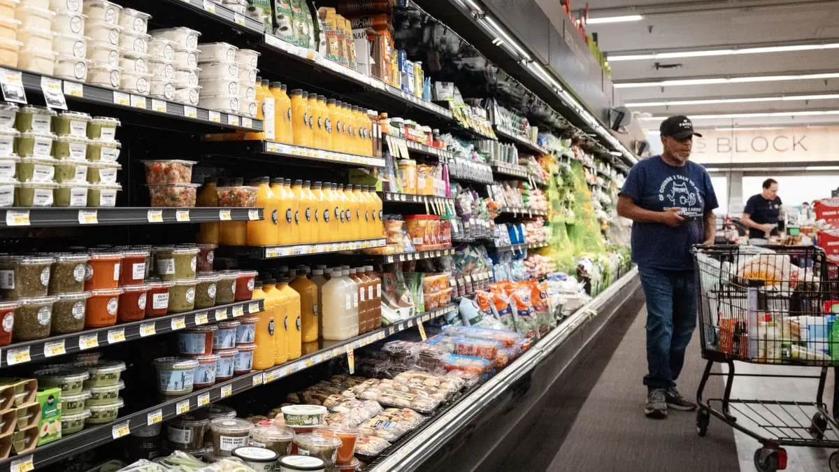Shoppers walk past refrigerated grocery shelves stocked with juice, produce, and prepared foods in a supermarket.