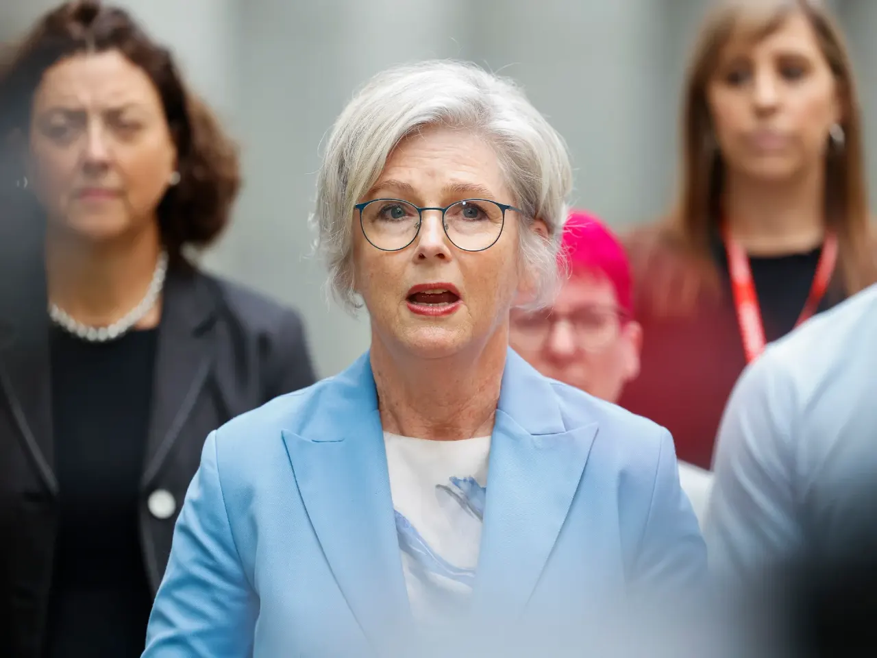 Silver-haired woman in blue blazer speaking, with two women blurred behind her