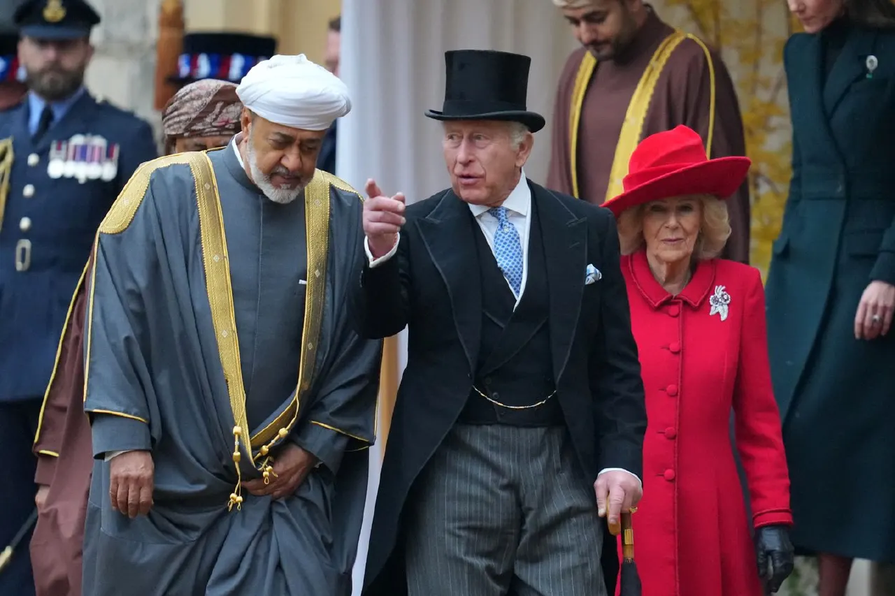 Formally dressed men and a woman in red walk together during a ceremonial visit, with uniformed attendants nearby.