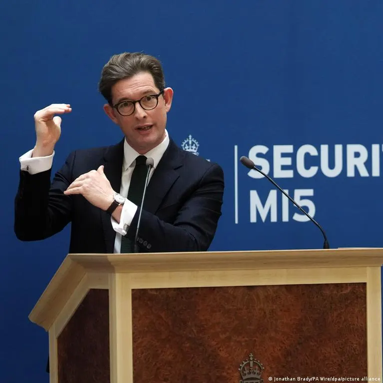 Man in dark suit and glasses speaking at podium before blue MI5 Security Service backdrop.