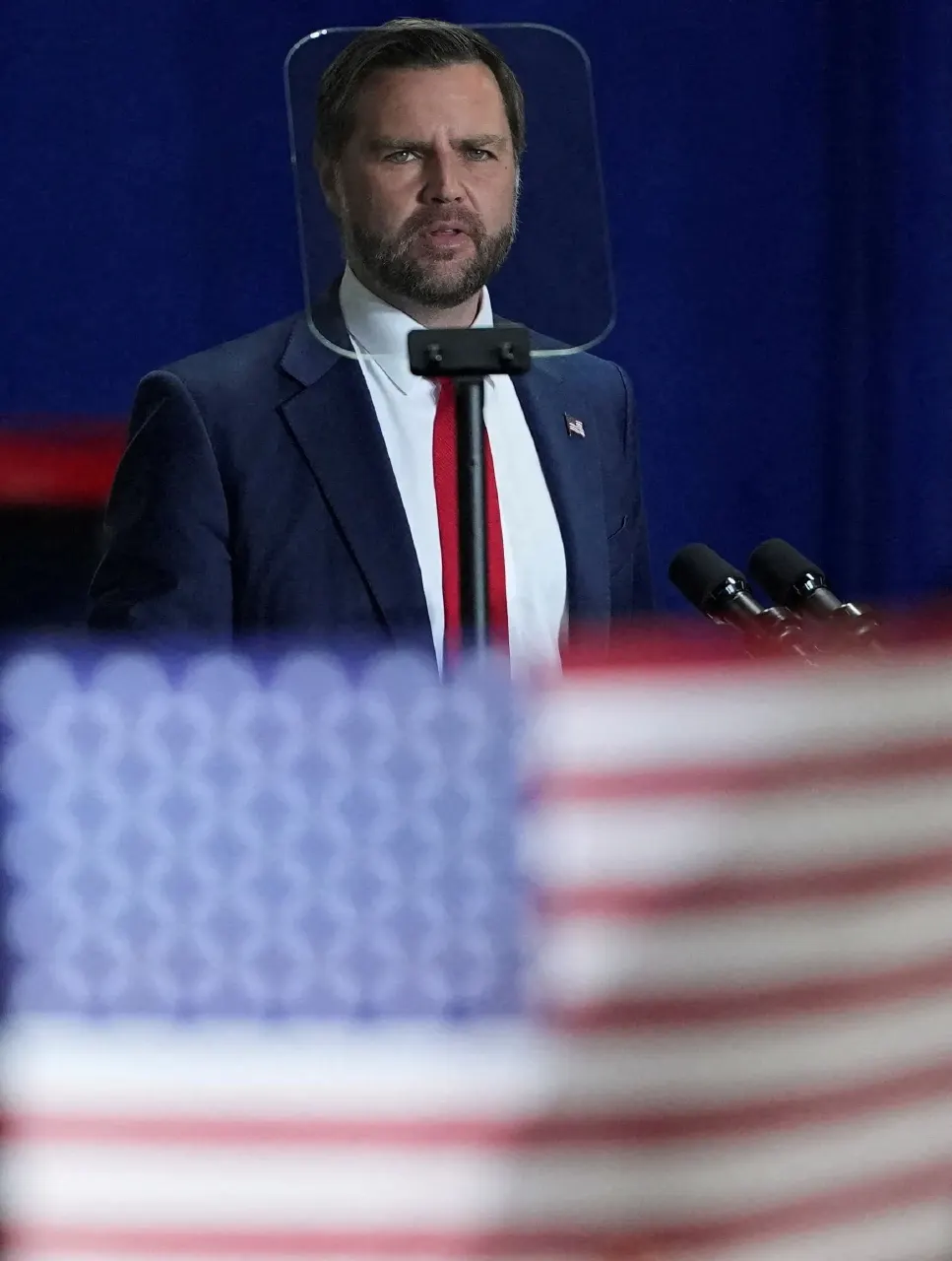 A man in a navy suit and red tie speaks at a podium behind a glass shield, with an American flag blurred in the foreground.