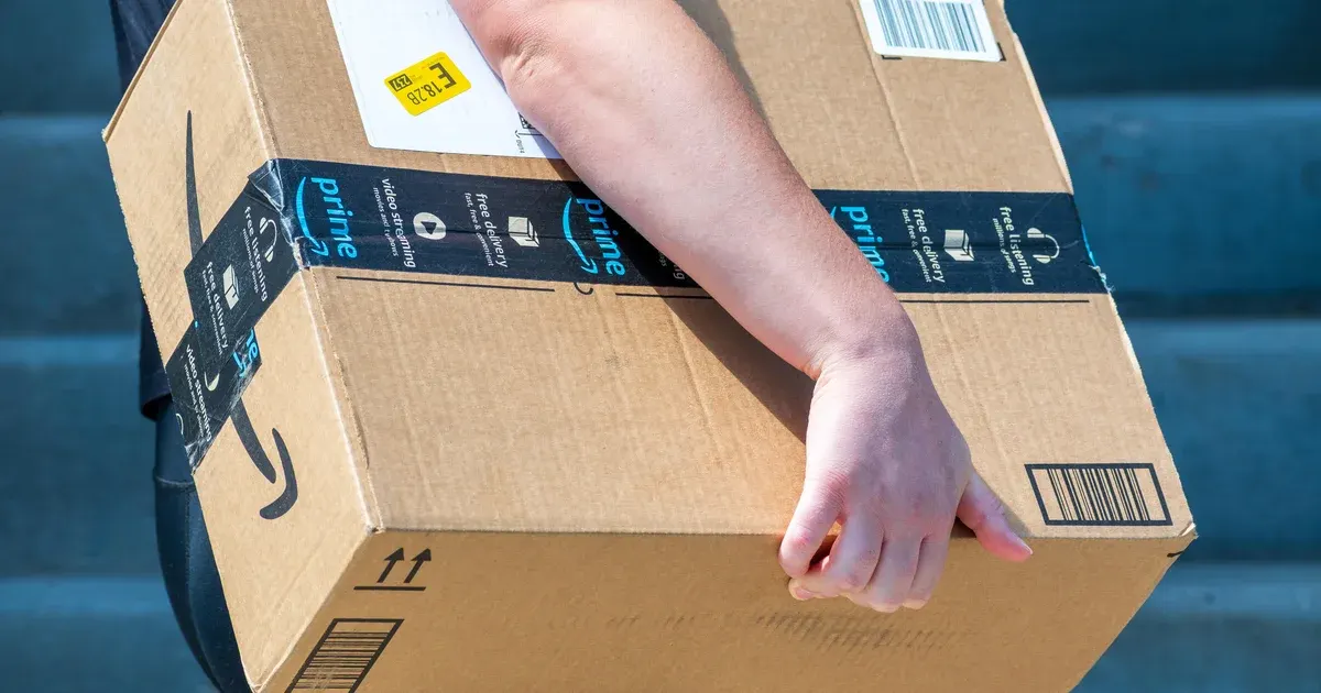 A person's arm carries a cardboard box with Amazon Prime tape and smile logo.