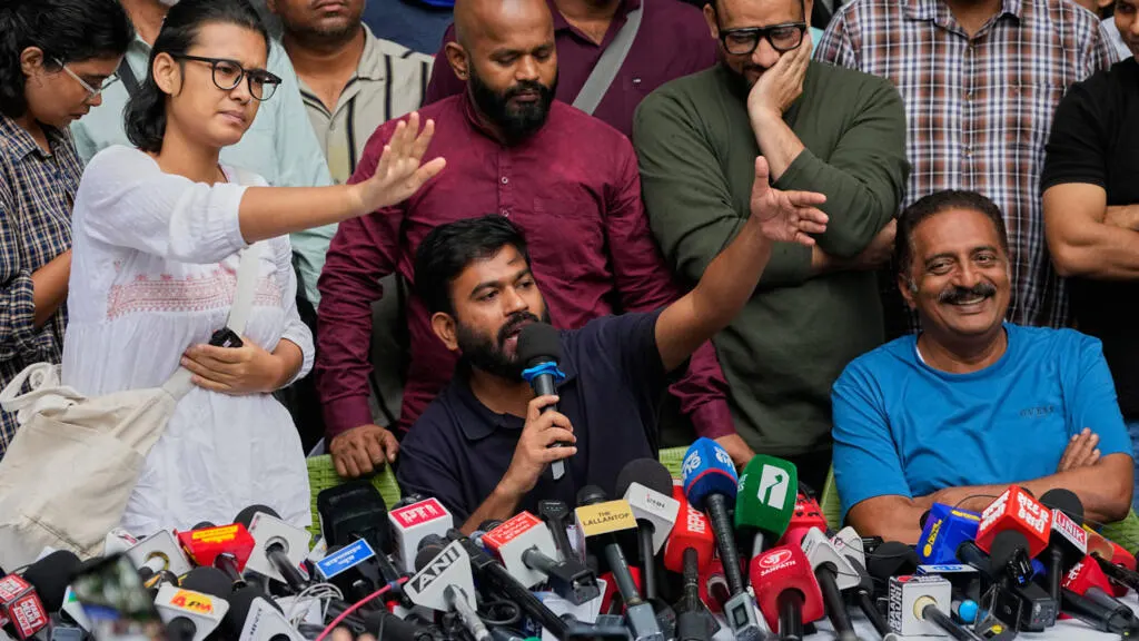 Bearded man speaks into microphone at press conference, surrounded by media mics and onlookers.