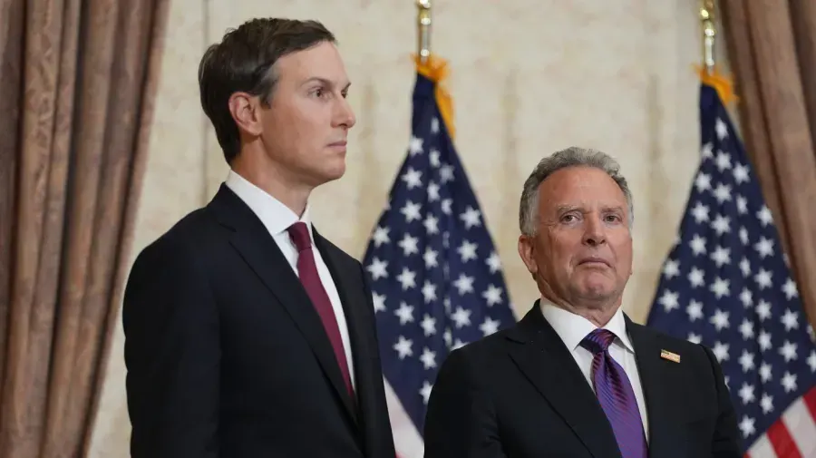 Two men in dark suits stand before American flags in a formal government setting.