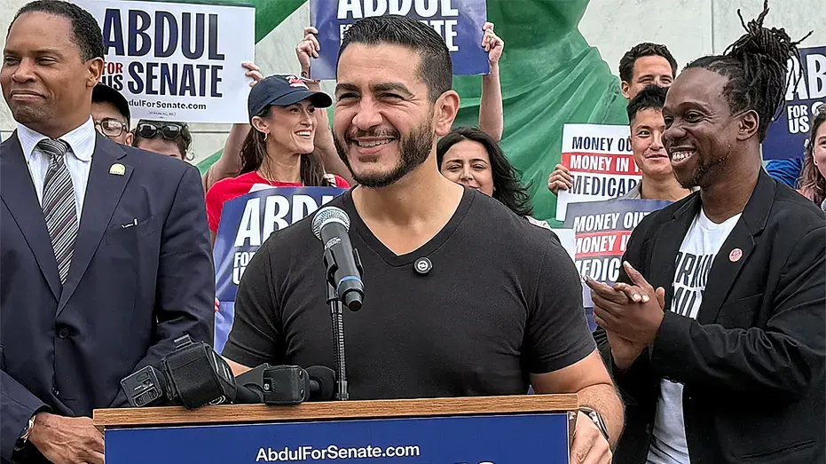 Man speaks at podium with campaign signs reading Abdul for Senate; supporters stand behind him.