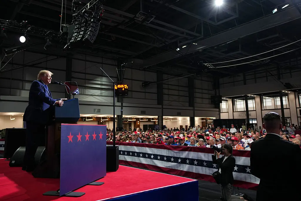 Speaker at a podium on a red stage before a large crowd, with a US flag banner.