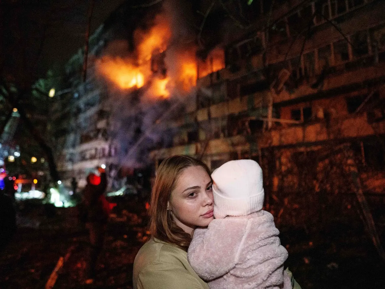 Woman holding a young child in front of a burning apartment building at night.