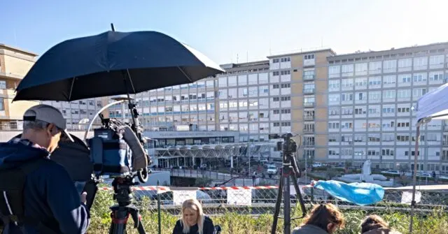 Camera crew with umbrella filming a large hospital building, with reporters waiting behind barrier tape.