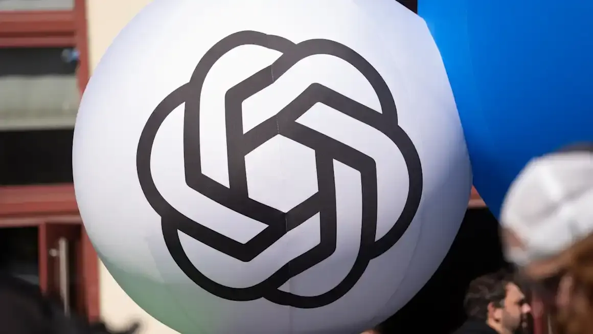 White inflatable ball with black OpenAI logo held outdoors, people blurred behind.