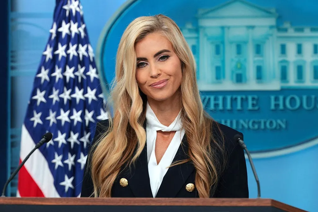 A woman with long blonde hair smiles at a podium before a White House seal and flag.