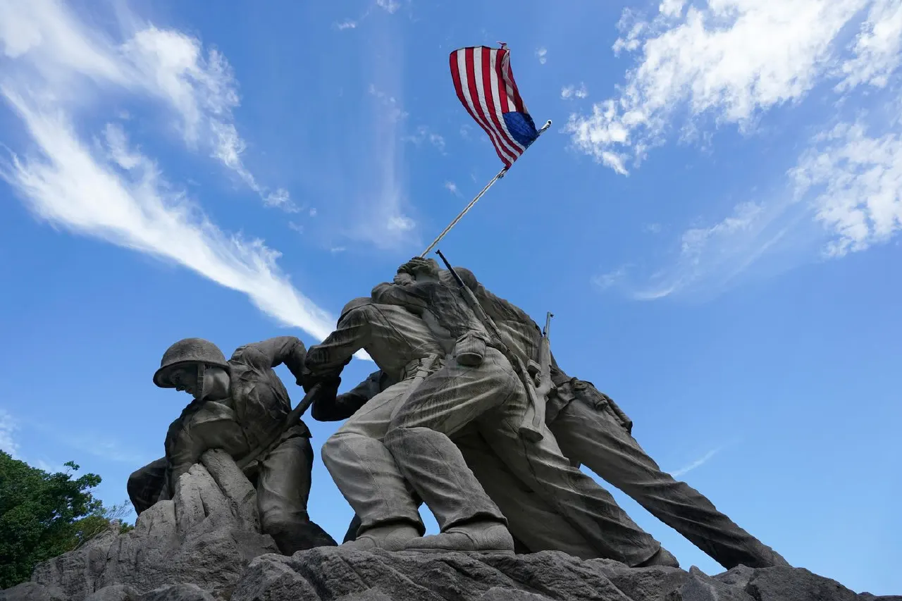 Bronze Iwo Jima flag-raising statue under a blue sky, with an American flag, shot from below.
