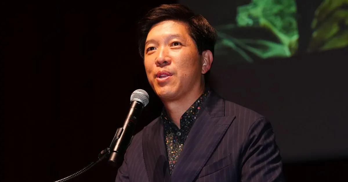 Man in pinstriped suit speaking at a microphone before a green projected screen.