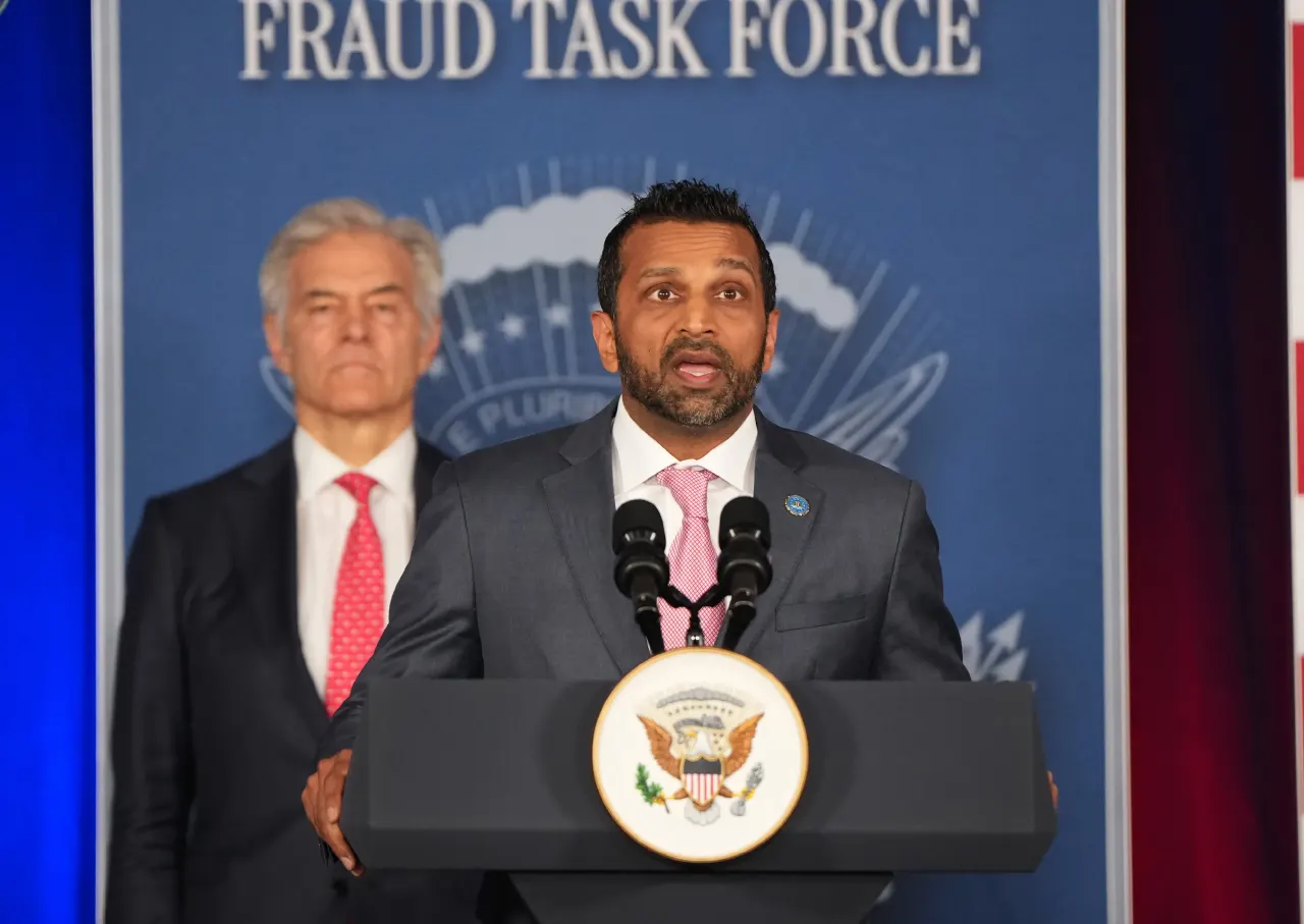 Official speaking at a podium with a presidential seal before a Fraud Task Force backdrop, another man behind.