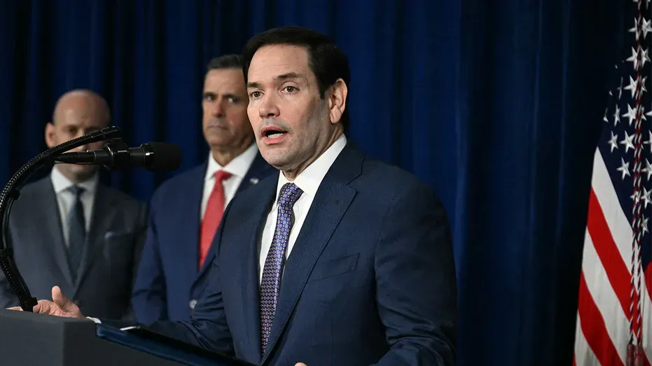 Man in dark suit speaking at a podium, American flag and blue curtain behind him.