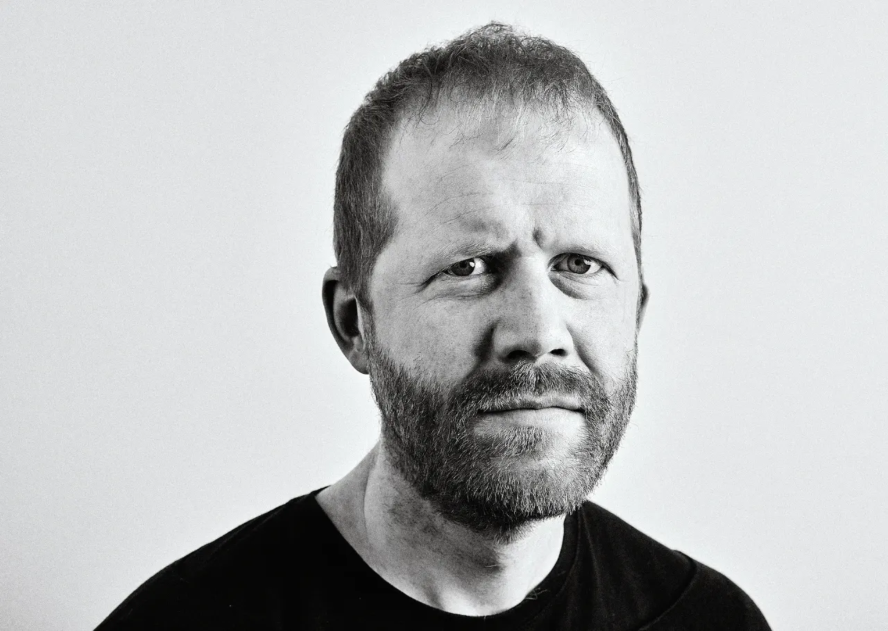 Black and white studio portrait of a bearded man in a dark shirt against a plain background.