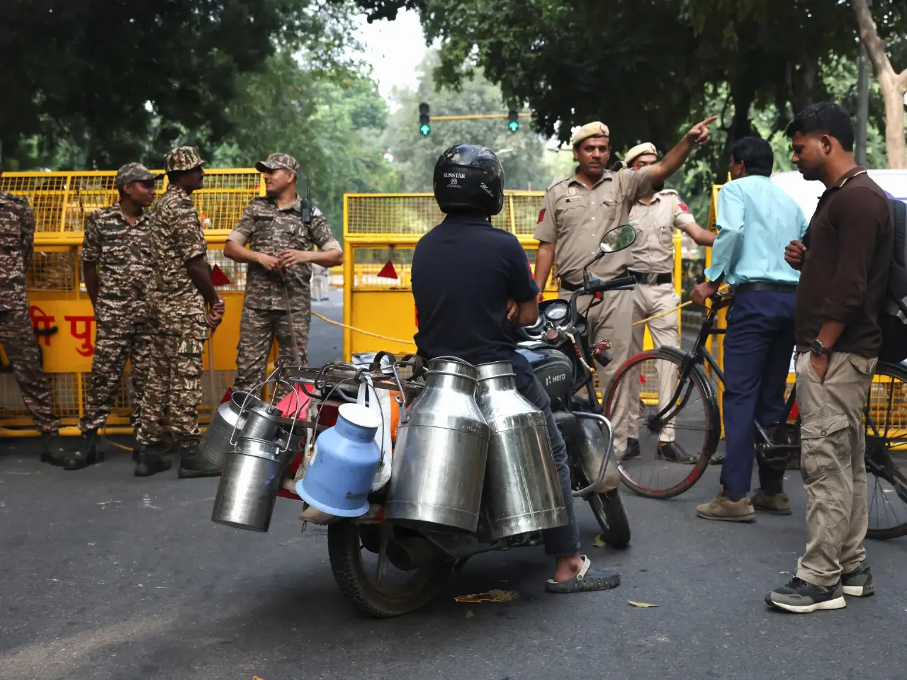 Camouflage-uniformed security personnel and police block a street; a motorcyclist with milk cans passes by.