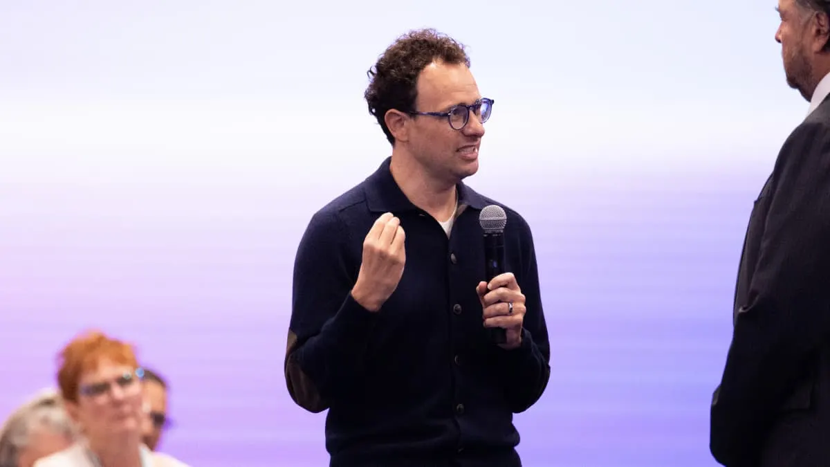 Man in glasses and dark cardigan speaking into microphone at a event, another person beside him.