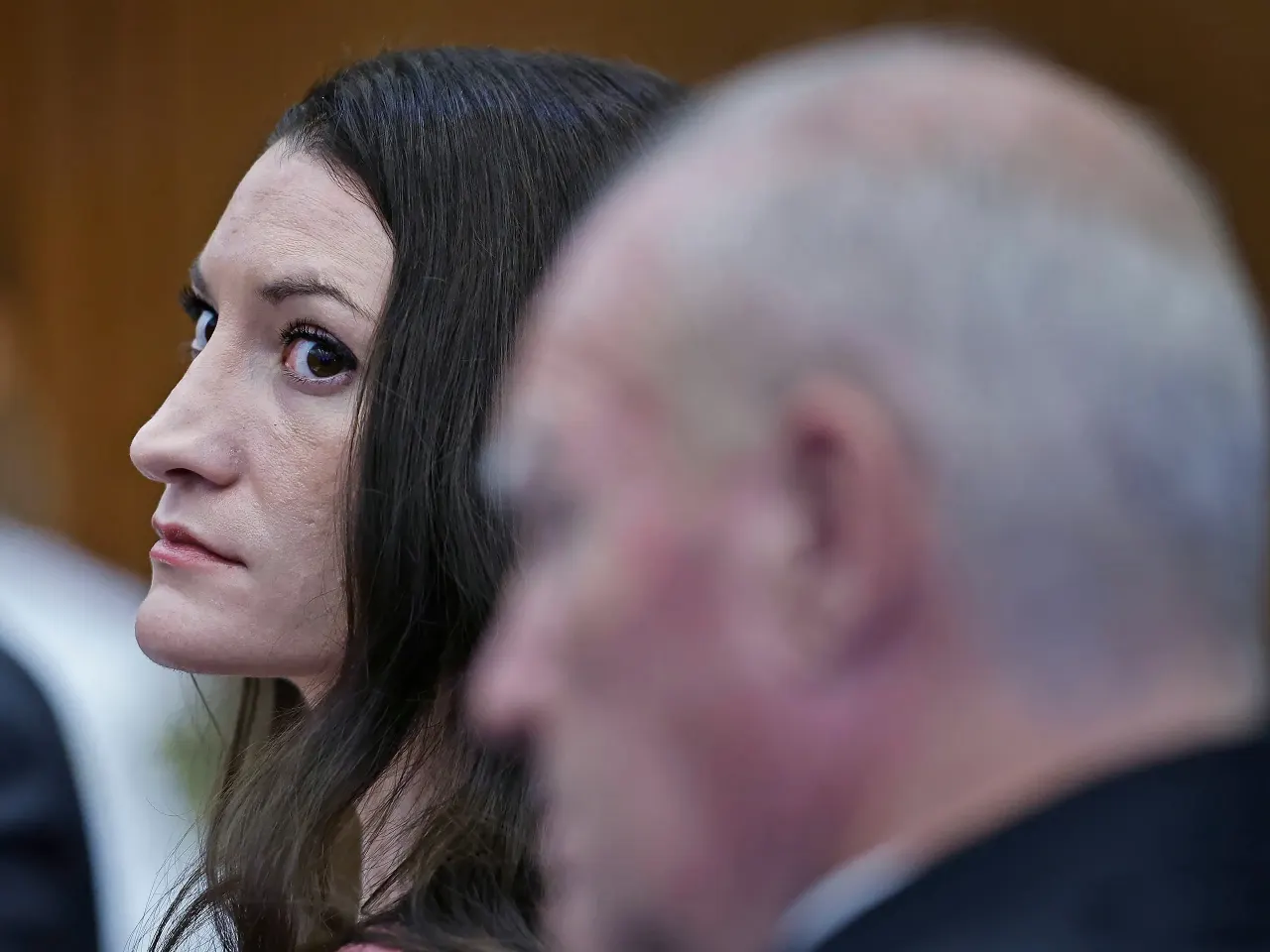 Woman with long dark hair looks sideways beside a blurred bald man in a courtroom setting.