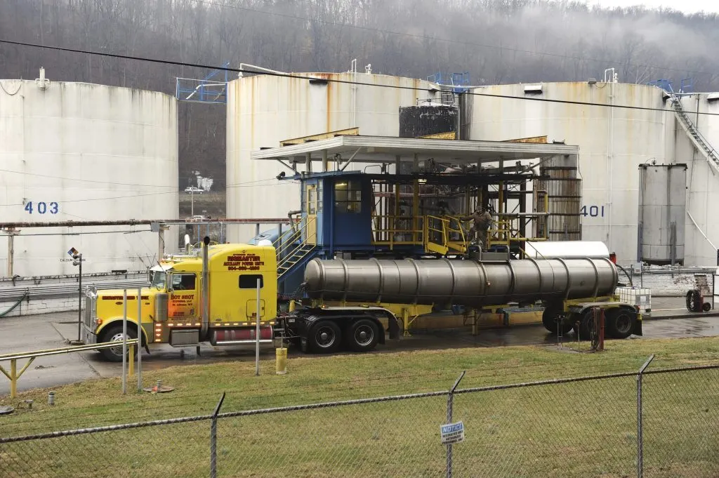 Yellow tanker truck with tank trailer parked beside large white industrial storage tanks at a terminal.