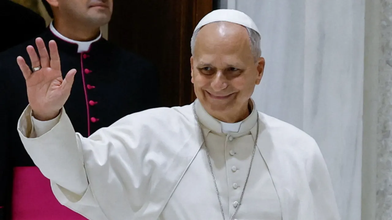 Pope in white cassock and zucchetto smiling and waving, with a clergyman behind him.