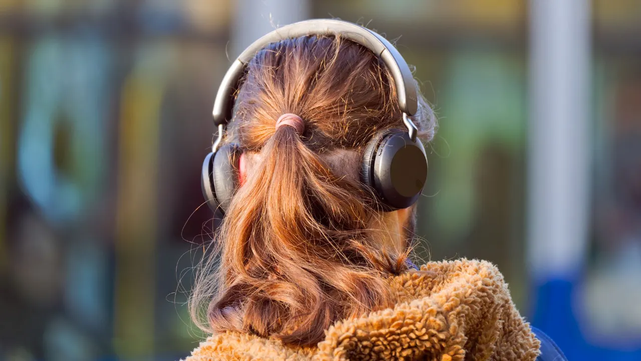 Back of a woman with headphones and ponytail, wearing a fuzzy tan jacket, blurred street background.