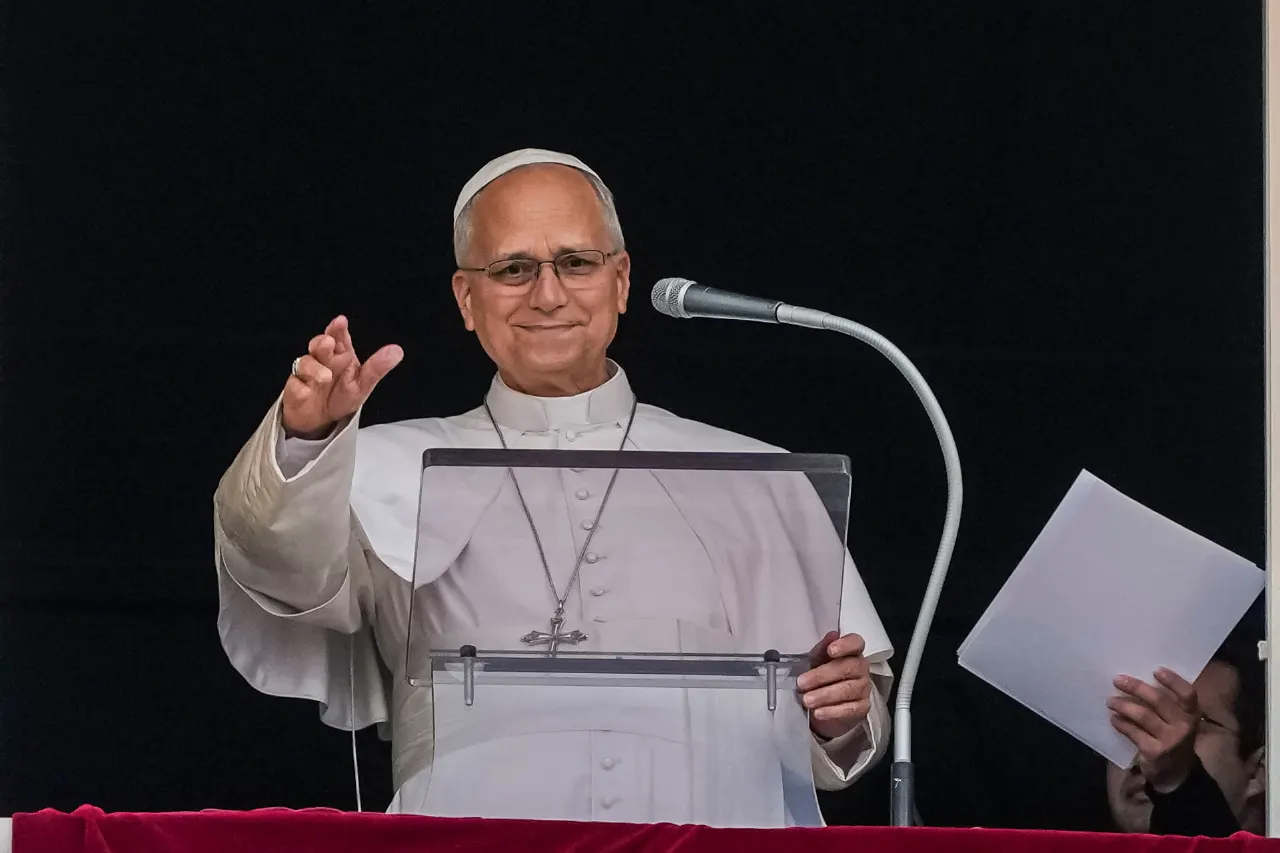 Man in white papal vestments and zucchetto smiling and gesturing at a lectern with microphone.