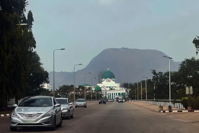 Street in Abuja with cars, green-domed government building and Aso Rock mountain behind.