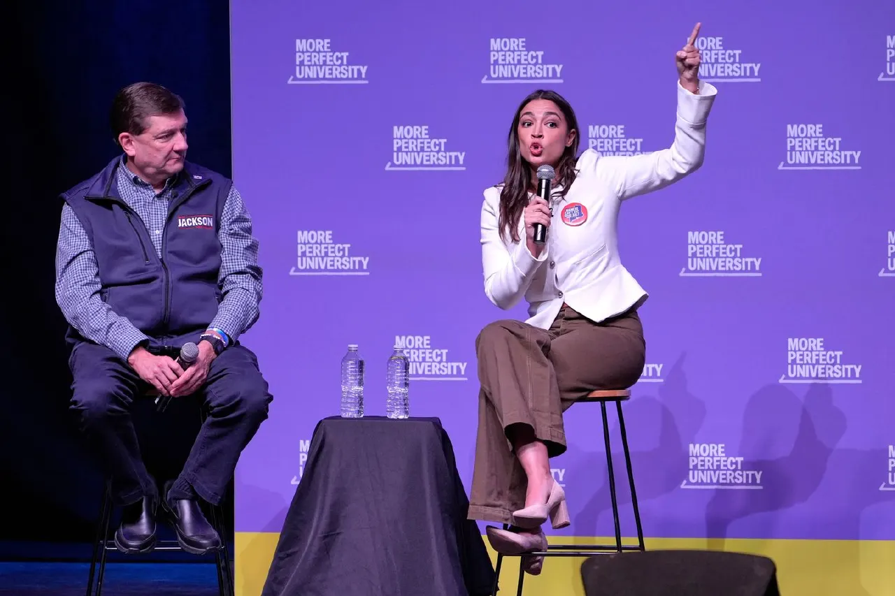 Two speakers on stage at a More Perfect University event; one holds a mic and raises a finger.