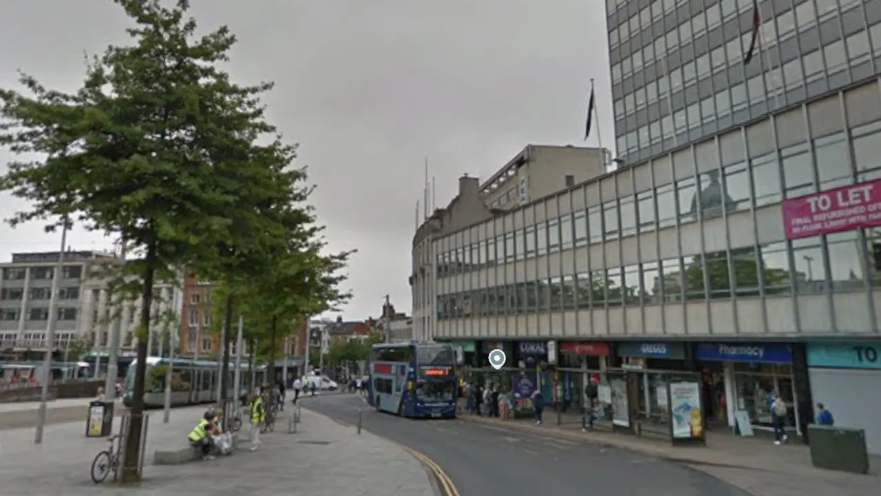 Street view of a city centre road with a bus, shops and a 'To Let' building.