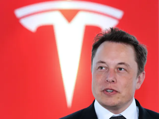 A man in a dark suit speaks in front of a large red Tesla logo.