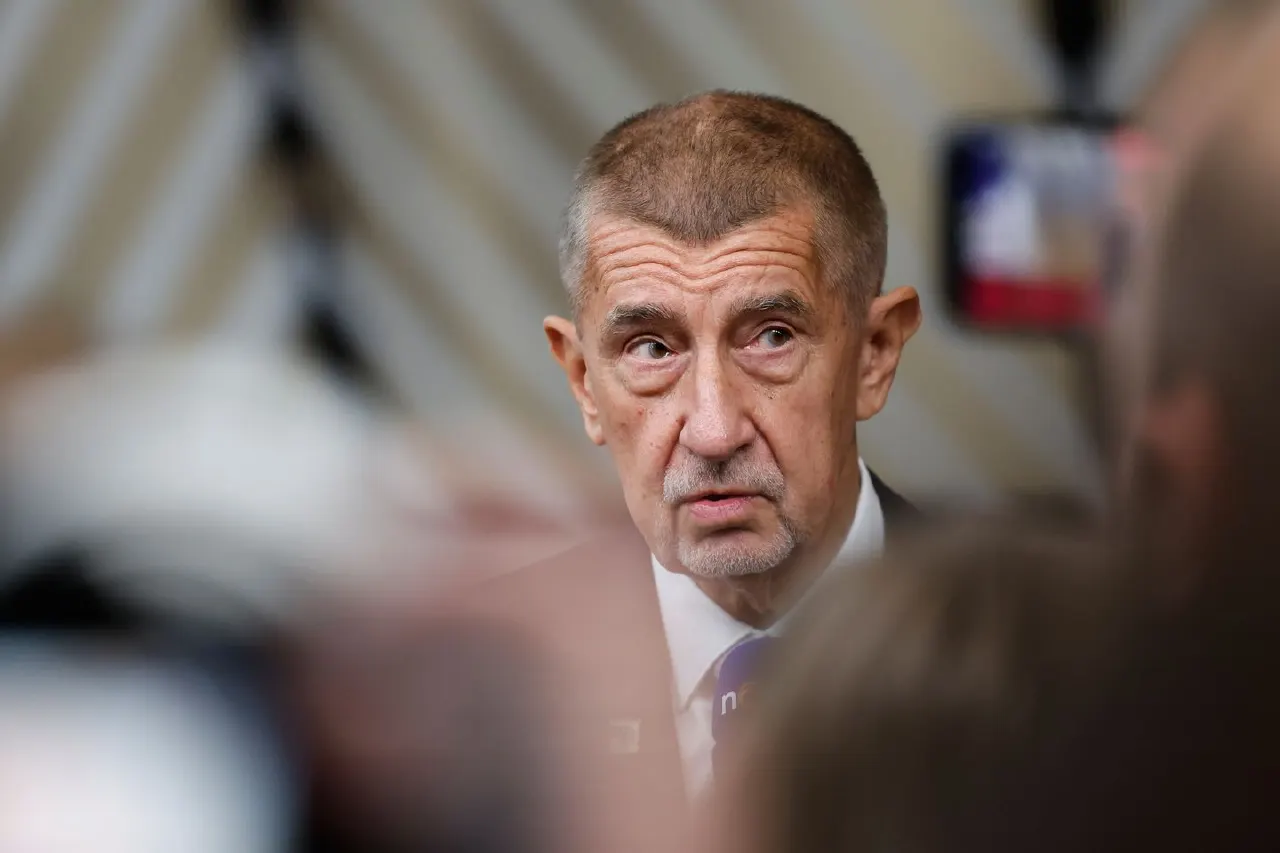 Older man with short grey hair speaking, surrounded by blurred figures and a camera in the background.