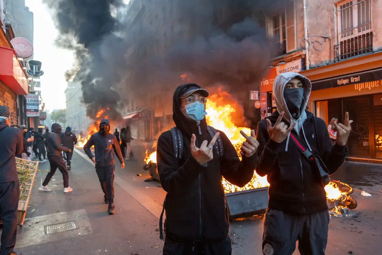 Masked, hooded youths gesture in a street with burning bins and black smoke behind them.