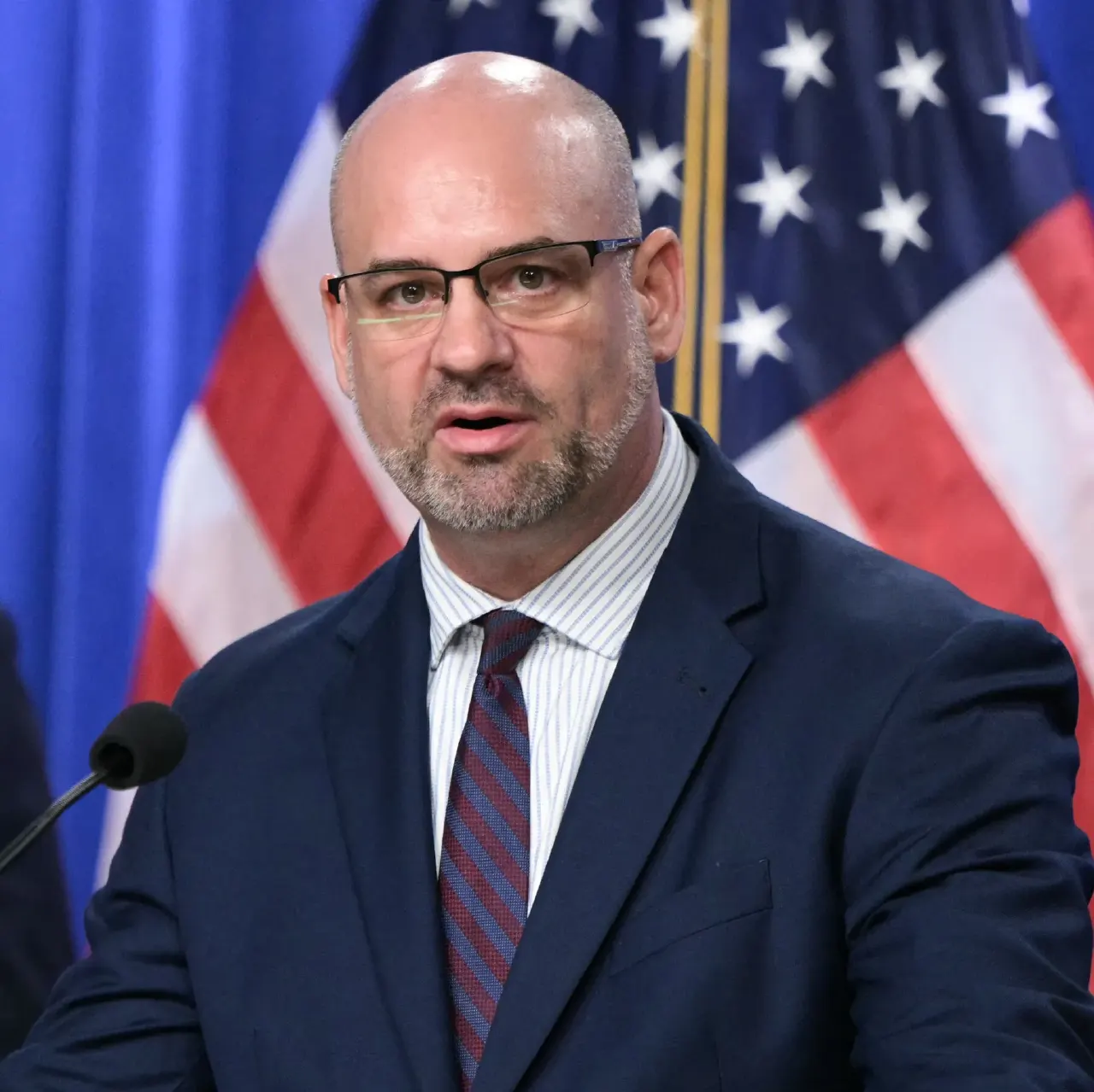 Bald man with glasses speaking at a podium before American flag and blue backdrop.
