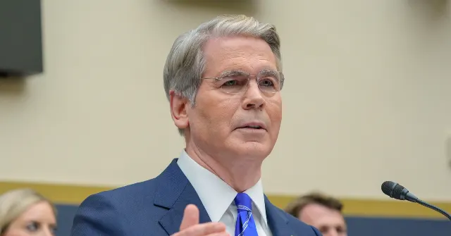 Man in blue suit and glasses speaking at a hearing microphone, gesturing with hand.