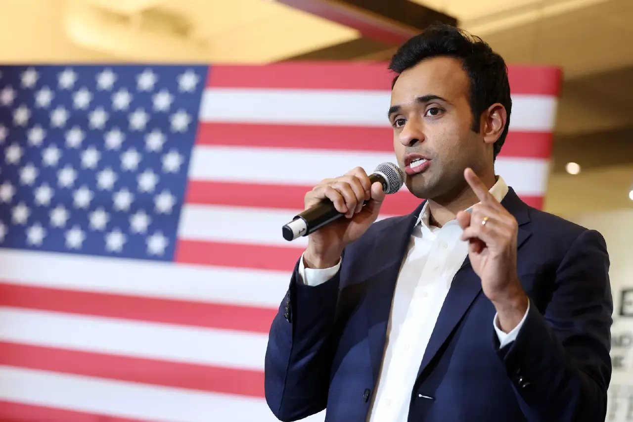 A man in a navy blazer speaks into a handheld microphone, pointing upward, in front of a U.S. flag.